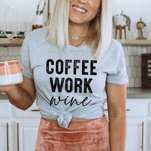 May include: A woman wearing a grey t-shirt with the text "COFFEE WORK wine" in black. She is holding a pink and white mug.