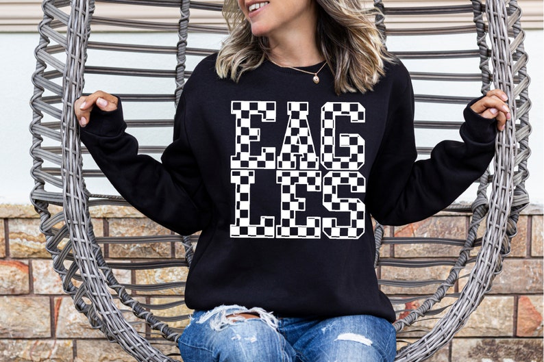May include: Black sweatshirt with the word "EAGLES" in large, checkered letters. The sweatshirt is a crew neck style and is being worn by a person. The background includes a woven chair and a brick wall.