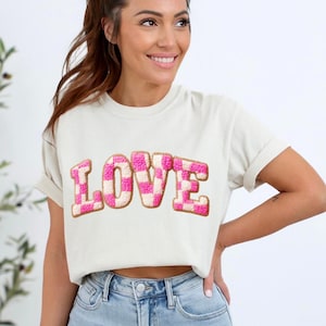 May include: Cream-colored t-shirt with the word "LOVE" in large, pink and white checkered letters. The letters have a textured, embroidered appearance, and the shirt has rolled-up sleeves.