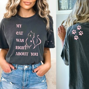 May include: A dark gray t-shirt with the text "My cat was right about you" in pink lettering. The shirt also features a pink outline of a cat with a heart on its chest. The back of the shirt has three pink paw prints.