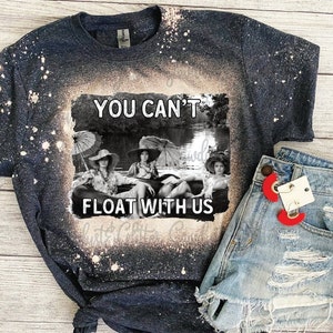 May include: A dark blue t-shirt with a bleach-splattered design featuring a black and white photo of three women in a boat with the text "You Can't Float With Us".