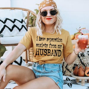 May include: Mustard yellow t-shirt with the text "I have separation anxiety from my HUSBAND". The person is wearing sunglasses and a patterned bandana. A glass of red liquid with a straw is held in the hand. Fruits are on a small wooden table.