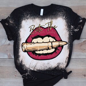 May include: A black t-shirt with a bleached design of a red mouth biting a gold bullet. The text "Bite The" is written in gold glitter above the mouth.