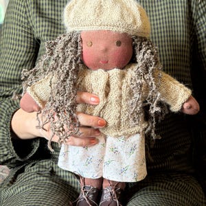 Può includere: Una bambola di stoffa fatta a mano con pelle scura, capelli castani ricci e tratti del viso ricamati. La bambola indossa un basco in maglia beige, un cardigan in maglia beige abbinato, pantaloncini bianchi con una piccola stampa floreale viola e scarpe marroni stringate.