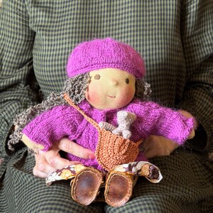 Può includere: Una bambola di stoffa fatta a mano con capelli castani ricci, che indossa un basco in maglia magenta e un maglione magenta abbinato. La bambola ha una piccola borsa a tracolla beige contenente un minuscolo giocattolo e scarpe marroni.