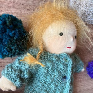 May include: A handmade fabric doll with messy ginger hair, wearing a textured sage green knitted cardigan with two buttons. The doll features simple embroidered facial details, including small black eyes and a thin red mouth, and is surrounded by colorful yarn pom-poms in shades of teal, purple, and white.
