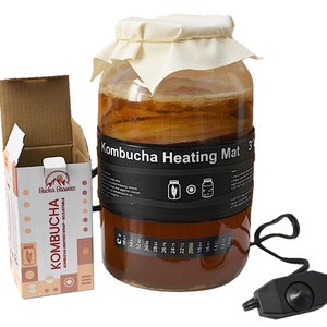 May include: A clear glass jar with a brown liquid inside, a white cloth covering the top, and a black heating mat wrapped around the middle. The heating mat has a temperature gauge and a switch. The jar is labeled "Kombucha Heating Mat 3". The jar is sitting on a white surface. The image also shows a cardboard box with the text "Bucha Brewers Kombucha Kombucha Heating Wrap - Adjustable" printed on it.