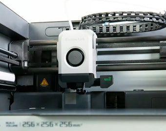 May include: Close-up of a 3D printer with a white extruder head and a black and gray build plate. The printer has a black and gray frame and a black belt.