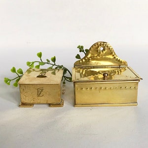 Pode incluir: Duas caixas de lat&atilde;o, uma com tampa articulada e a outra com uma tampa pequena e quadrada. A caixa com a tampa articulada tem um design decorativo na frente e um pequeno bot&atilde;o no topo. A caixa menor tem a letra "E" gravada na frente.
