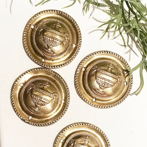 Könnte beinhalten: Vier goldfarbene Metall-Schubladengriffe mit verzierten Designs, die eine stilisierte Urne mit einem floralen Motiv zeigen. Die Griffe haben einen perlenbesetzten Rand und ein leicht erhabenes Design.