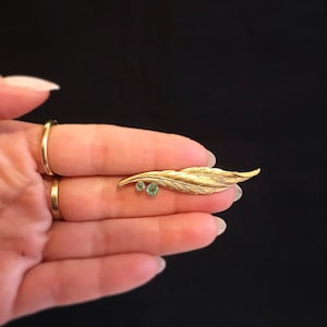 Könnte beinhalten: Eine goldfarbene Federbrosche mit zwei grünen Edelsteinen. Die Brosche wird in einer Hand gehalten.
