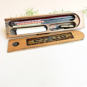 May include: A wooden pencil box with a decorative inlay on the lid. The box is open and contains a variety of pencils, including graphite, colored, and a white eraser.