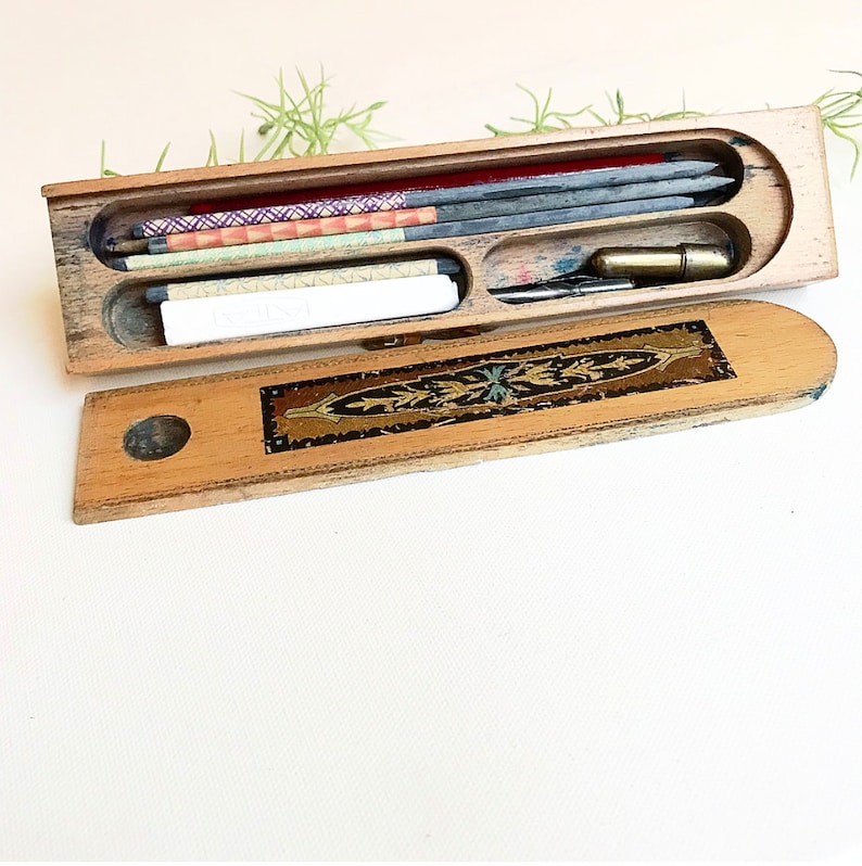 May include: A wooden pencil box with a decorative inlay on the lid. The box is open and contains a variety of pencils, including graphite, colored, and a white eraser.