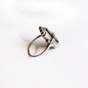 May include: A silver ring with a black stone and a small pearl accent. The ring has an intricate design and is set in a geometric shape.