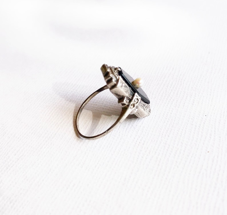 May include: A silver ring with a black stone and a small pearl accent. The ring has an intricate design and is set in a geometric shape.