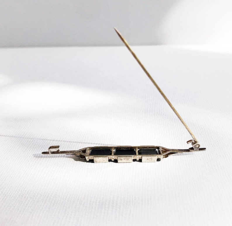 May include: A silver brooch with a row of black rectangular stones. The brooch has a pin and catch closure.