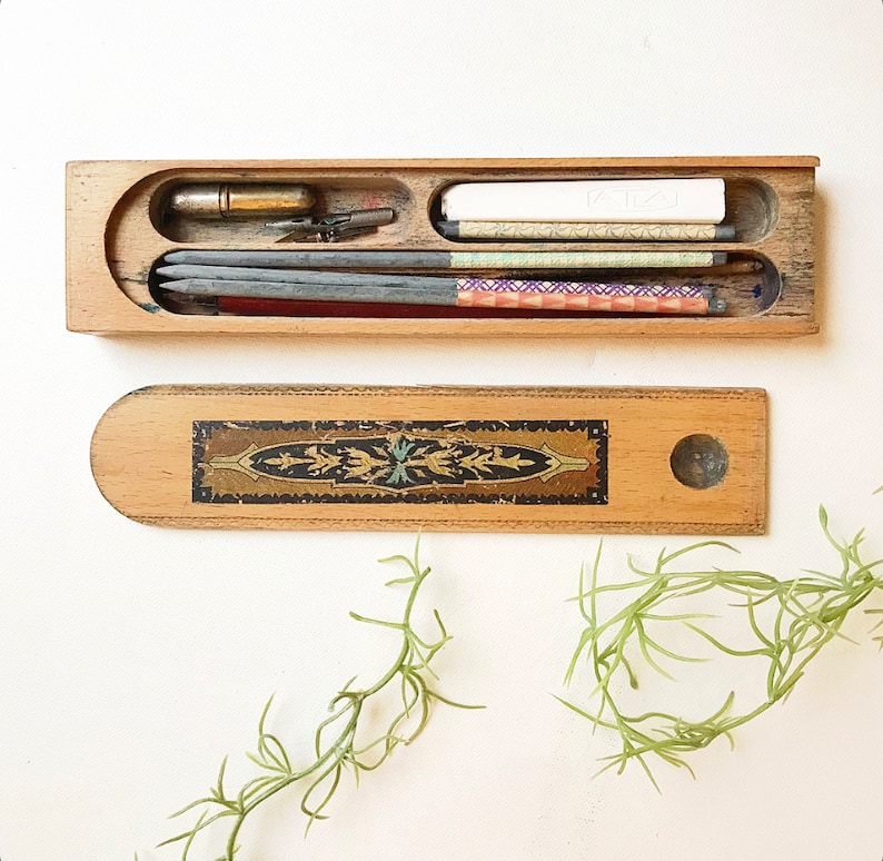 May include: A wooden pencil box with a decorative inlay. The box is open and contains a variety of pencils, a ruler, and a small eraser. The box is made of light wood with a dark brown inlay.