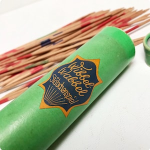 Puede incluir: Un tubo verde con una etiqueta dorada y azul que dice "Wibbel Wabbel Stäbchenspiel!" El tubo contiene un juego de palitos de madera con puntas rojas, azules y verdes.