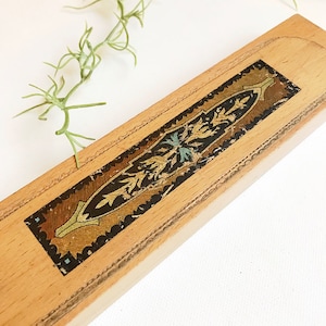 May include: A wooden pencil box with a decorative, ornate design in gold, black, and blue. The box is made of light wood and has a rectangular shape.