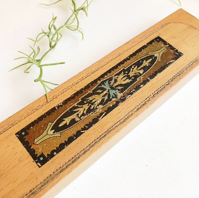 May include: A wooden pencil box with a decorative, ornate design in gold, black, and blue. The box is made of light wood and has a rectangular shape.