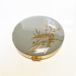 Può includere: Un compatto rotondo di colore grigio con un bordo dorato e un motivo floreale in oro e marrone.