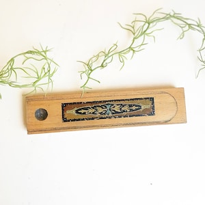 May include: A wooden pencil box with a decorative inlay of gold and blue floral patterns on a black background. The box has a round hole on the side.