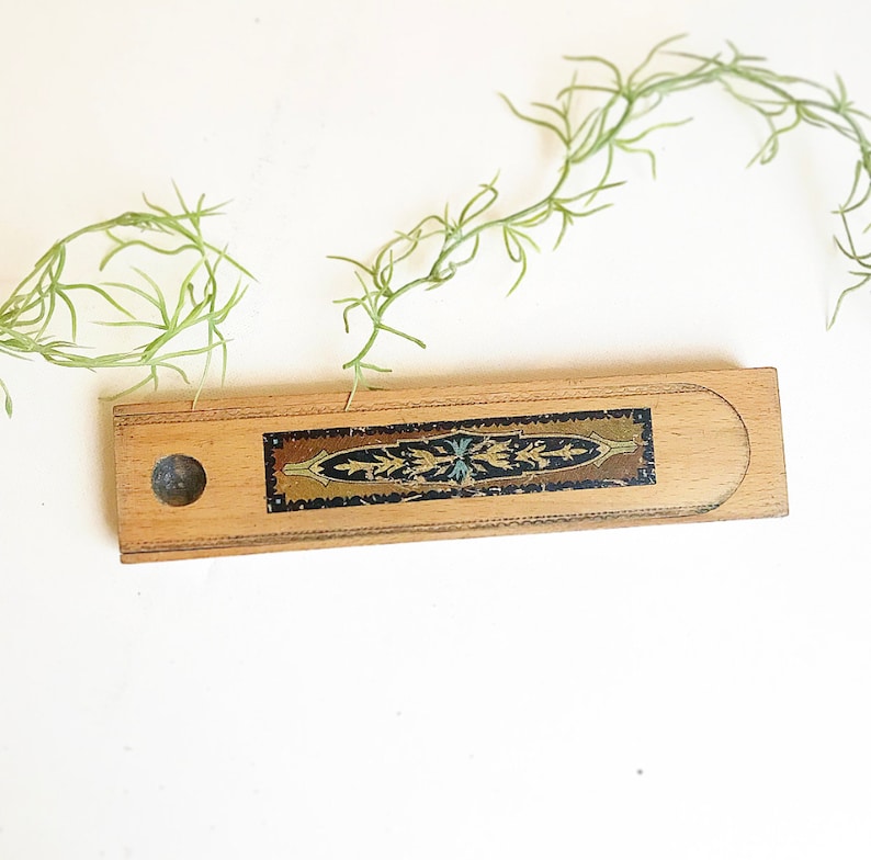 May include: A wooden pencil box with a decorative inlay of gold and blue floral patterns on a black background. The box has a round hole on the side.