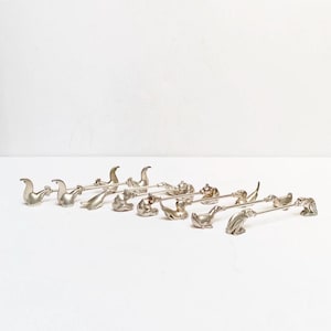 Pode incluir: Um conjunto de garfos de coquetel prateados em forma de animais. Cada garfo apresenta uma figura de animal diferente, incluindo focas, pássaros e outras criaturas. Os garfos estão dispostos em duas fileiras sobre um fundo branco.