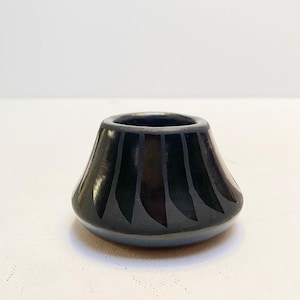 Può includere: Un piccolo vaso in ceramica nera con forma svasata e ampia apertura. Il vaso presenta una serie di linee verticali, curve e di colore chiaro che creano un motivo a ventaglio. Il vaso è appoggiato su una superficie bianca.