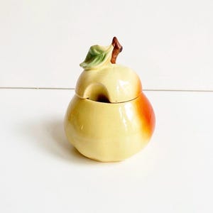 Può includere: Un contenitore in ceramica a forma di mela con coperchio. Il contenitore è principalmente giallo con una sfumatura rosso-arancio su un lato. Il coperchio presenta una foglia verde e un gambo marrone. Il contenitore è probabilmente per conservare alimenti.