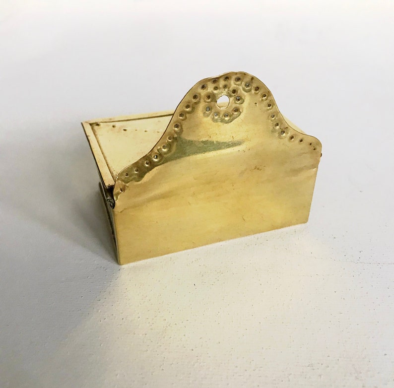Pode incluir: Um pequeno porta-palitos de f&oacute;sforo de lat&atilde;o com uma parte superior decorativa festoneada. O porta-palitos tem uma tampa com dobradi&ccedil;as e uma base retangular.