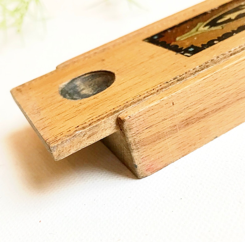 May include: A wooden box with a hinged lid and a small round hole in the top. The box has a decorative design on the top with a gold and blue pattern.