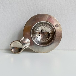 Peut inclure: Une passoire à thé argentée avec un bol rond perforé et une poignée. La passoire a une petite poignée décorative et un petit emblème sur le bord. La passoire à thé est conçue pour être placée sur une tasse pour retenir les feuilles de thé.
