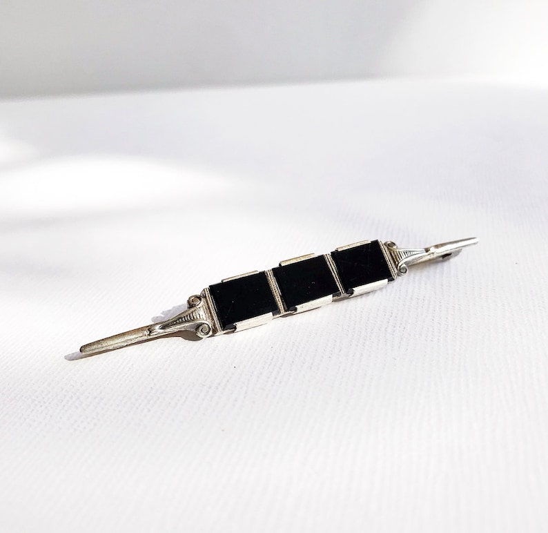 May include: A silver brooch with three black square stones set in a row. The brooch has a simple, elegant design.