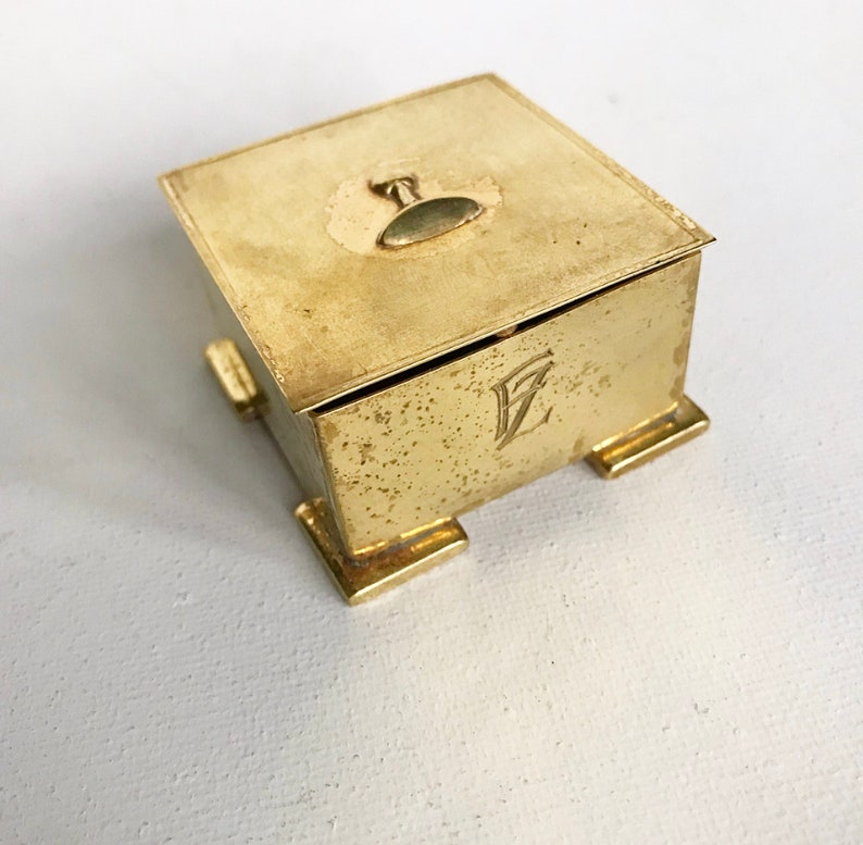Pode incluir: Uma pequena caixa quadrada de lat&atilde;o com uma tampa com dobradi&ccedil;as e um pequeno bot&atilde;o redondo no topo. A caixa tem quatro p&eacute;s e &eacute; gravada com a letra "Z".