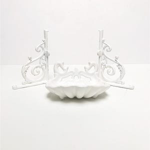 Pode incluir: Suportes de prateleira de metal branco com um design decorativo de volutas e um saboneteira correspondente em forma de concha de mar de metal branco.