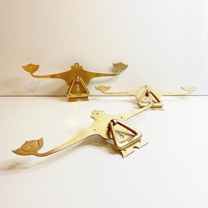 Könnte beinhalten: Drei messingfarbene dekorative Griffe mit Vintage-Ästhetik. Jeder Griff hat einen zentralen dreieckigen Griff und stilisierte, flügelartige Verlängerungen. Die Griffe sind auf einer weißen Oberfläche angeordnet und zeigen ihr kompliziertes Design und ihre warme Metalloberfläche.