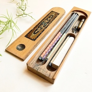 May include: A wooden pencil box with a decorative inlay on the lid. The box is open and contains several pencils, a pencil sharpener, and a white eraser. The pencils are wrapped in colorful paper.