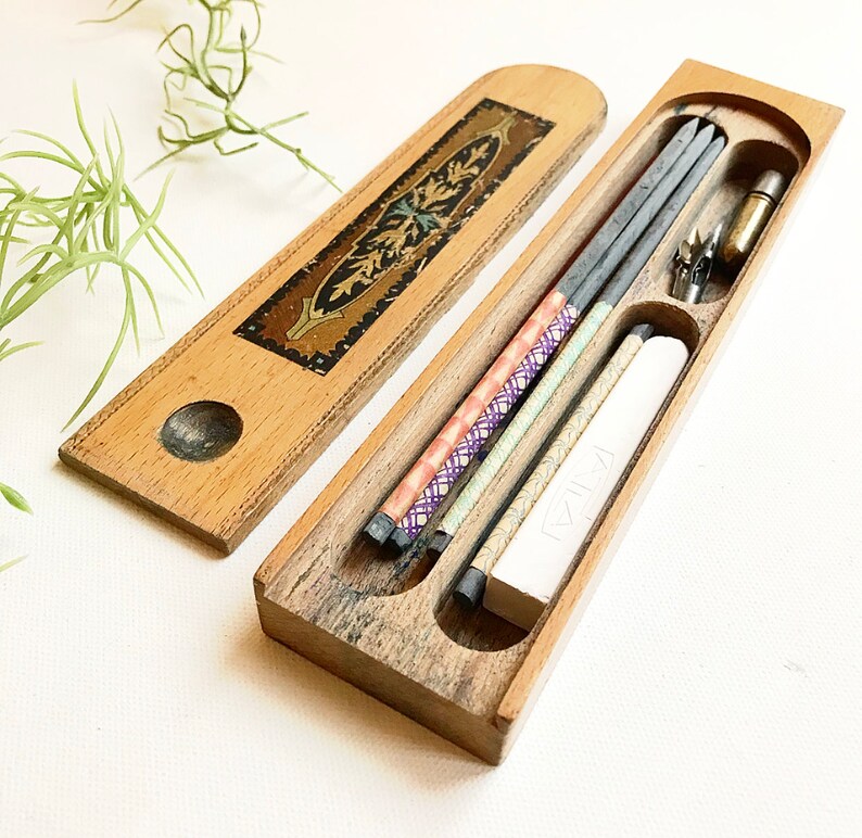 May include: A wooden pencil box with a decorative inlay on the lid. The box is open and contains several pencils, a pencil sharpener, and a white eraser. The pencils are wrapped in colorful paper.