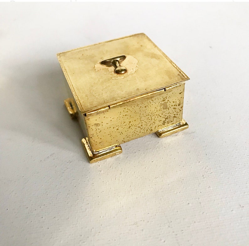 Pode incluir: Uma pequena caixa quadrada de metal de cor dourada com uma tampa articulada e um pequeno bot&atilde;o redondo no topo. A caixa tem uma superf&iacute;cie texturizada e assenta em quatro pequenos p&eacute;s.