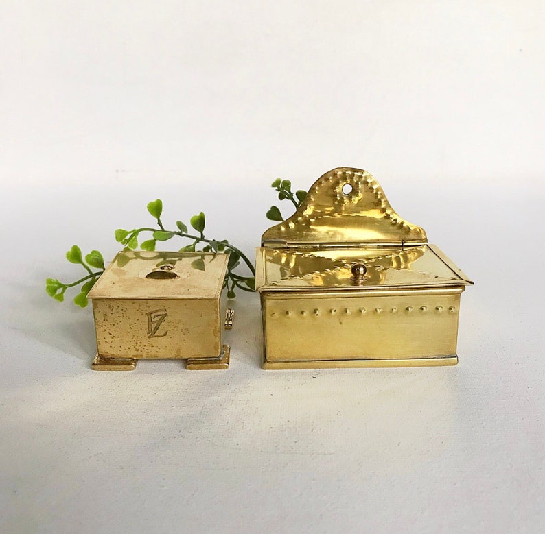 Pode incluir: Duas caixas de lat&atilde;o, uma com tampa articulada e a outra com uma tampa pequena e quadrada. A caixa com a tampa articulada tem um design decorativo na frente e um pequeno bot&atilde;o no topo. A caixa menor tem a letra "E" gravada na frente.