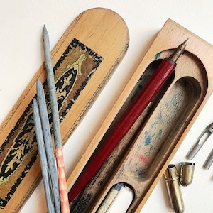 May include: A wooden box with a hinged lid, containing a red pen, several gray pencils, and a few metal nibs. The box has a decorative design with gold accents.