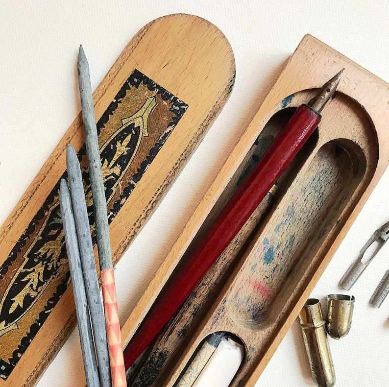 May include: A wooden box with a hinged lid, containing a red pen, several gray pencils, and a few metal nibs. The box has a decorative design with gold accents.