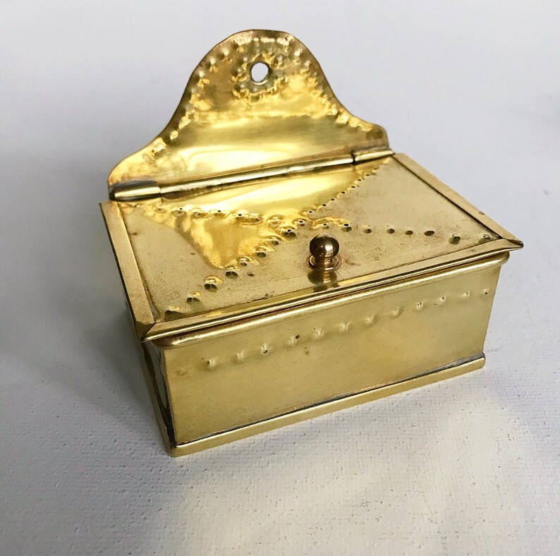 Pode incluir: Uma pequena caixa de lat&atilde;o antiga com tampa articulada e bot&atilde;o decorativo. A caixa tem uma superf&iacute;cie texturizada e uma borda festonada.