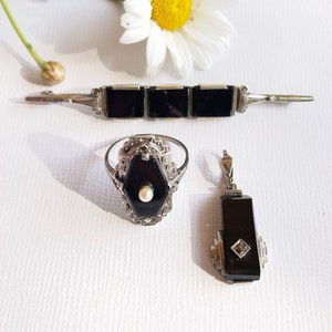 May include: A set of three vintage jewelry pieces featuring black onyx and silver. The set includes a bracelet, a ring, and a pendant. Each piece is adorned with intricate filigree details and a single pearl accent.