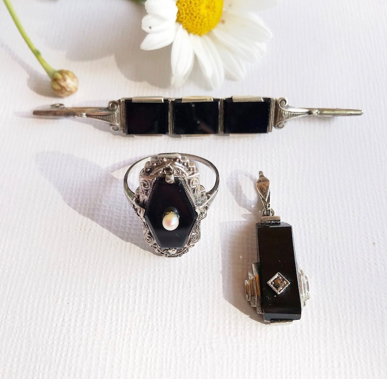May include: A set of three vintage jewelry pieces featuring black onyx and silver. The set includes a bracelet, a ring, and a pendant. Each piece is adorned with intricate filigree details and a single pearl accent.