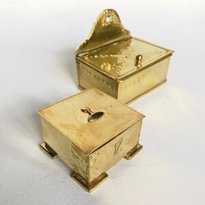 Pode incluir: Duas caixas antigas de lat&atilde;o. A caixa maior tem uma tampa com dobradi&ccedil;as e um design decorativo. A caixa menor &eacute; quadrada e tem quatro p&eacute;s. Ambas as caixas t&ecirc;m um bot&atilde;o de lat&atilde;o na parte superior.