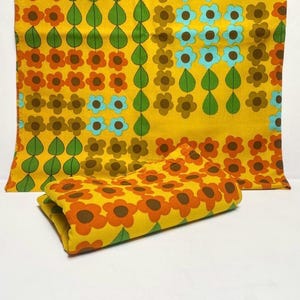 Può includere: Un tessuto giallo piegato con un motivo floreale ripetuto in arancione, marrone e azzurro. Il design include file di foglie verdi. Il tessuto è probabilmente un tessuto vintage o un oggetto decorativo.