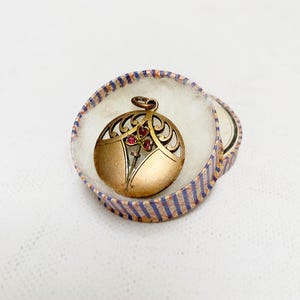 Op de afbeelding: Een goudkleurige hanger met een opengewerkt ontwerp, met een bloemmotief met rode accenten, rust in een gestreepte doos. De hanger is rond en heeft een kleine lus voor een ketting. De doos heeft blauwe en oranje strepen.