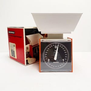 May include: A vintage SOEHNLE kitchen scale with a white weighing bowl and a black dial. The dial features white numerals and markings, with a needle pointing to zero. The scale has an orange frame and comes with its original red box.